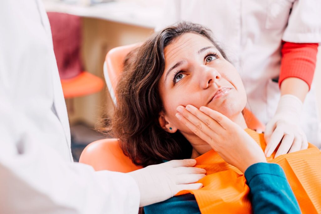 Woman in dental chair holding hand to face in pain looking at dentist